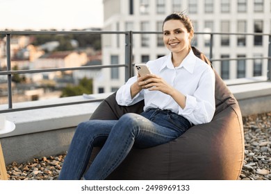 Woman relaxing on a rooftop beanbag chair using smartphone. She is smiling, wear white shirt, denim jeans, enjoying outdoor space with urban background. - Powered by Shutterstock