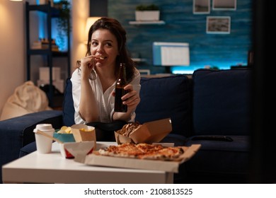 Woman relaxing on sofa watching television while drinking craft beer and eating potato chips in home living room. Office worker enjoying a cold beer and tasty home deliverd fast food.