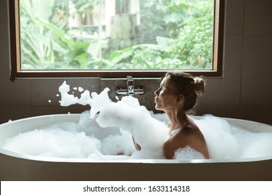 Woman Relaxing With Pleasue In Foam Bath With Bubbles In Modern Hotel Dark Bathroom By Big Window With Tropical View