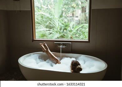 Woman Relaxing With Pleasue In Foam Bath With Bubbles In Modern Hotel Dark Bathroom By Big Window With Tropical View