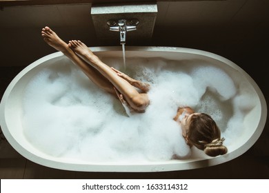 Woman relaxing with pleasure in foam bath with bubbles and touching her smooth leg skin in modern hotel dark bathroom, top view