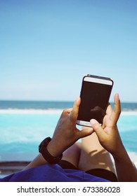 Woman Relaxing Pool Bed Or Sundeck On Beach Lounge Swimming Pool Sea View For Vacation Using A Mobile Phone Near The Pool. User POV. Female Holding Mobile Phone.with Copy Space. 