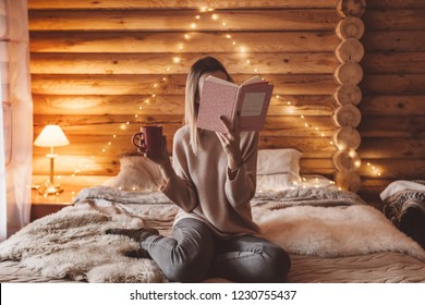 Woman relaxing and reading book on cozy bed in log cabin in winter