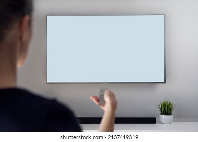 A Woman With Remote Control Sitting On Couch And Watching TV At Home (Smart TV), Space For Design On Screen. Business, Logo, Free Space To Use. Artificial Grass And Soundbar In The Background.