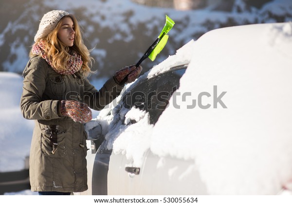 Woman remove snow\
from car with snow brush.