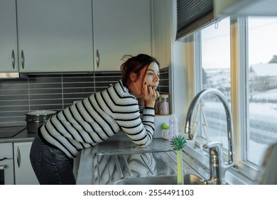 Woman resting her chin in her hand, gazing pensively at a snowy scene through the kitchen window on a winter day - Powered by Shutterstock