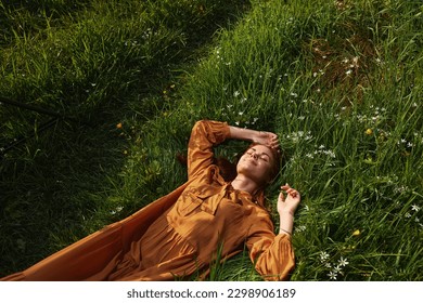  woman, resting lying in the green grass, in a long orange dress, with her eyes closed and a pleasant smile on her face, holding her hands near her face, enjoying harmony with nature and recuperating - Powered by Shutterstock