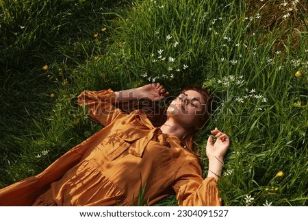  woman, resting lying in the green grass, in a long orange dress, with her eyes closed and a pleasant smile on her face, holding her hands near her face, enjoying harmony with nature and recuperating