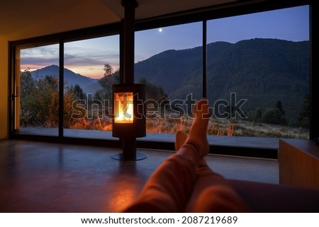 Woman resting near fireplace at modern house with great view on mountains at sunset. Women's legs on background of landscape