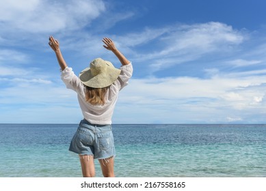 A woman is resting on the beach. She is wearing a straw hat, denim shorts and a white shirt. Travel and good rest, happy holidays, tourism, Philippines, summer season. Rear view.