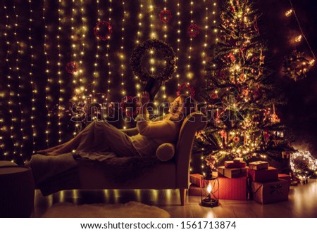 Woman resting on couch in dark and reading a book on Christmas Eve, decorated room on background. Vintage and long exposure effect. Christmas magic concept.