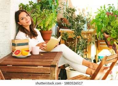 Woman resting on cozy balcony with many potted plants, reading a book