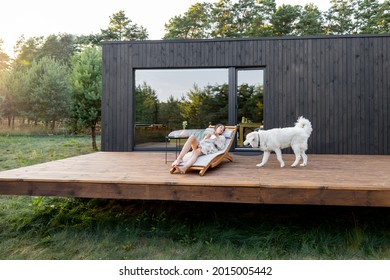 Woman resting on sunbed on the wooden terrace near the modern house with panoramic windows near pine forest with big white dog. Wellness and mindful health. Copy space - Powered by Shutterstock