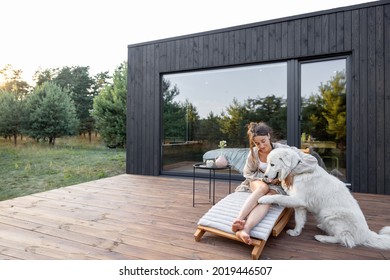 Woman resting on sunbed on wooden terrace near the modern house with panoramic windows near pine forest while playing with pet. Concept of solitude and recreation on nature