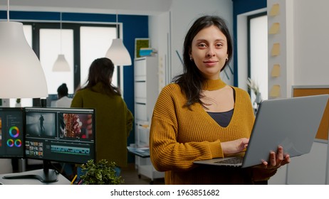 Woman retoucher looking at camera smiling working in creative media agency standing in front of webcam holding laptop. Photographer graphic editor working in start up office digital assets.