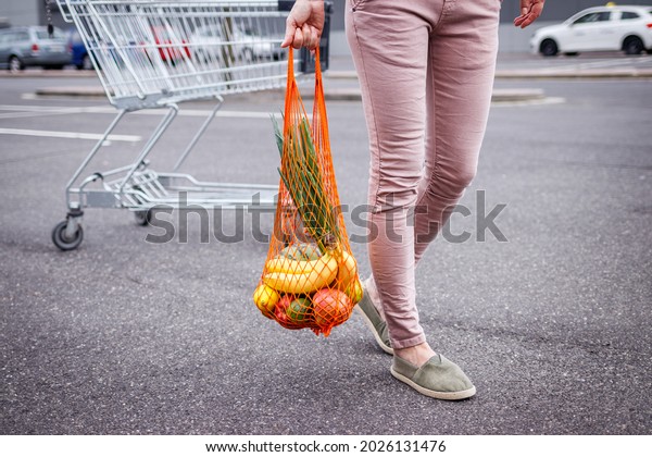 Woman with reusable mesh bag walking from\
shopping cart in parking lot next to supermarket. Plastic free and\
zero waste concept. Groceries\
purchase