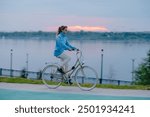 Woman rides a bicycle along a riverside path during sunset, enjoying a peaceful evening outdoors by the water.Evening cycling before going to bed, an active lifestyle