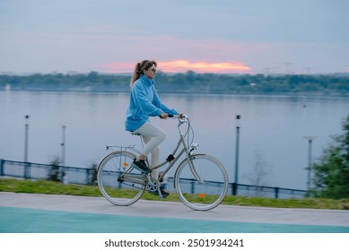 Woman rides a bicycle along a riverside path during sunset, enjoying a peaceful evening outdoors by the water.Evening cycling before going to bed, an active lifestyle - Powered by Shutterstock