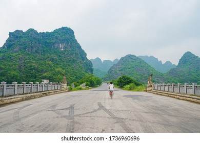 Woman Riding Bicycle Alone On The Road In Scenic Landscape Among Rock Pinnacles And Green Valleys. Unique View Of Trang An Ninh Binh, Vietnam. Social Distancing, Lone Traveling, Alternative Mobility.