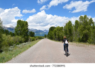 Woman Riding A Bicycle Along A Bike Path In Jackson Hole, Wyoming. The Grand Tetons Are In The Distance.