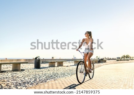 Woman riding bicycle on beach promenade in summer. Happy lady on bike having fun on seaside boulevard. Sunny waterfront street for cycling. Blue sky, summertime freedom. Carefree feeling at sunset.