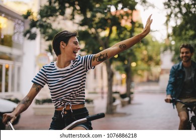 Woman Riding Bicycle And Waving Back To A Male Friend. Couple Enjoying Cycling Through City.