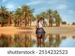 A woman riding a camel across the thin sand dunes sand dunes surround the oasis with palm tree and lake - Sahara, Morocco