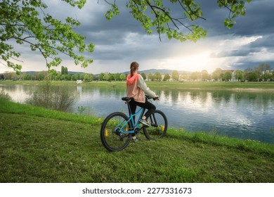 Woman riding a mountain bike near lake at sunset in spring. Colorful landscape with sporty girl, bicycle, coast of river, green grass, cloudy sky in park in summer. Sport and travel. Biking - Powered by Shutterstock