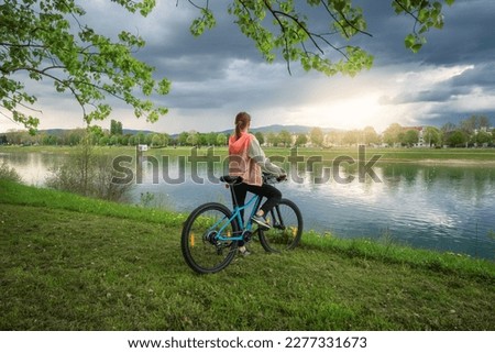 Woman riding a mountain bike near lake at sunset in spring. Colorful landscape with sporty girl, bicycle, coast of river, green grass, cloudy sky in park in summer. Sport and travel. Biking