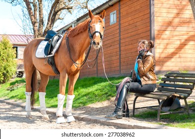 Woman Riding Trainer Near Chestnut Horse Speaking On Cell Phone. Multicolored Outdoors Image.