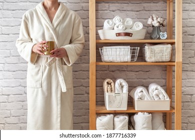 Woman In Robe Is Standing And Holding White Wire Basket Of Folded Bed Sheets Near Organized Linen Closet In Bathroom. Sorted And Folded Towels In White Wicker And Wire Baskets Placed On Wooden Shelves