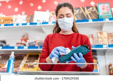 A Woman In Rubber Gloves And A Medical Mask Treats The Terminal With Antiseptic. In The Background Are Shelves Of The Store. The Concept Of Coronovirus And Sanitation