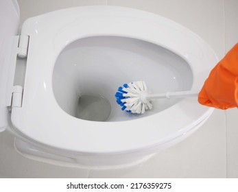 Woman with rubber orange glove is cleaning toilet bowl using brush.  Closeup photo, blurred. - Shutterstock ID 2176359275