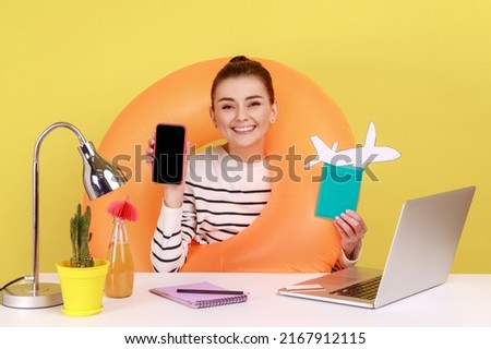 Woman with rubber ring, holding passport and paper plane, showing phone with empty display, advertisement of summer tours, mobile app. Indoor studio studio shot isolated on yellow background.