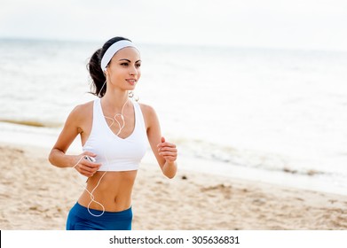 Woman Runner Running On The Beach Listening To Music On Smartphone Using Earphones. Female Fitness Girl Jogging On Path In Amazing Fall Foliage Landscape Nature Outside.