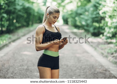 Woman Runner in the Summer Morning Park Listening to Music on Smartphone Using Bluetooth Earphones. Female Fitness Girl Jogging on Path Outside.