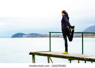 Woman Runner Warming Up By The Lake In Winter Time Preparing For Training. Stretching The Leg, Holding Her Running Shoe