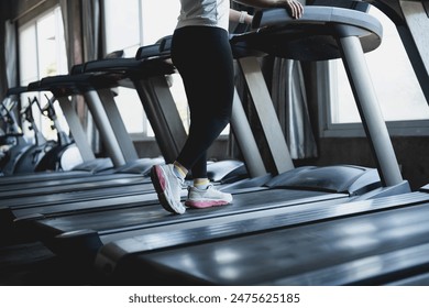 A woman is running on a treadmill in a gym. She is wearing white sneakers and black pants