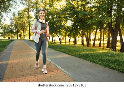 Woman running outdoors. Slim lady of senior age wearing wireless headphones on neck jogging on cobblestone trail in park. Short-haired female in activewear taking care of health by regular training.