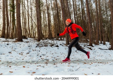 Woman Running Through Woods In Winter. Side View Of Smiling Female Runner Wearing Hydration Vest Trail Running In Forest On Cold Winter Day.