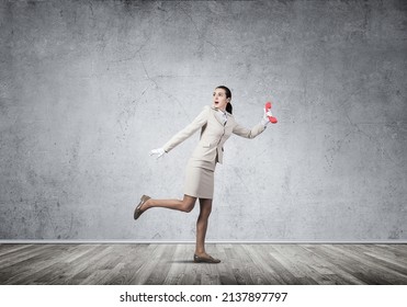 Woman Runs Away From Something With Vintage Red Phone. Call Center Operator In White Business Suit Posing With Telephone. Hotline Telemarketing And Communication. Professional Business Assistance.