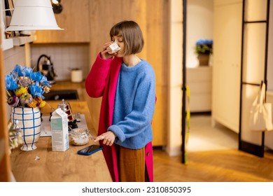 Woman rushes out of home putting on a coat, finishing her coffee and looking at smart phone. Concept of daily routine and busy work