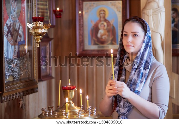 woman in the Russian Orthodox Church with red hair\
and a scarf on her head lights a candle and prays in front of the\
icon.