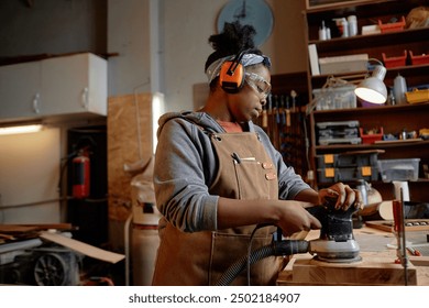 Woman sanding wood piece in organized woodworking space, wearing earmuffs and safety goggles for protection. Various tools and gadgets on shelves in background - Powered by Shutterstock