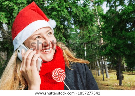 A woman in a Santa Claus hat stands in the park under a Christmas tree, has fun talking on a mobile phone and holds a lollipop in her hand. Christmas and New Year concept.