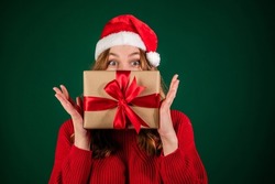 Woman In Santa Hat Covering Her Face With New Year Gift Isolated On Green Background. Smiling Girl In Sweater Peeks Over Christmas Box