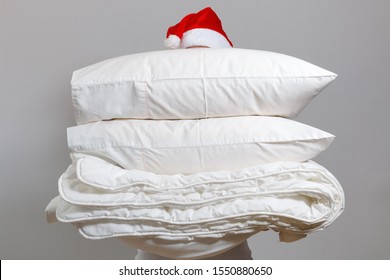 Woman In Santa Hat Holding A Warm Duvet And Feather Pillows Against A Gray Wall. Stack Of Bedding For Sleeping. Household At Christmas.