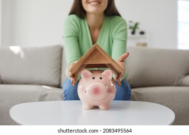 Woman Saving Up Money To Buy Her Own House. Happy Young Girl Holding Roof Above Small Pink Piggy Bank Placed On Table In Living Room, Close Up. Finance, Property Purchase, Taking Mortgage Loan Concept