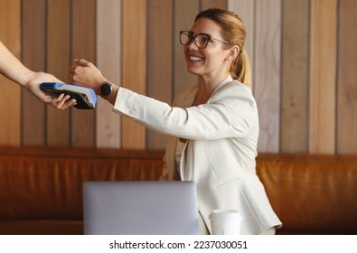 Woman scanning her smartwatch on a card machine to make a payment. Professional woman using NFC technology. Business woman buying coffee while working in a cafe. - Powered by Shutterstock