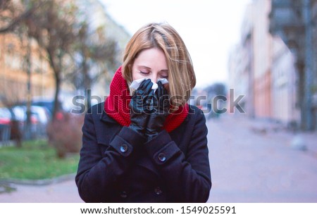 Woman with a scarf in her hands. the sick girl blows her nose in handkerchief. Seasonal colds and flu on a cloudy day. Sad blonde sneezes, outdoors or outside. On the street of the city, allergy.